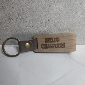 Könnte beinhalten: Ein Holzschlüsselanhänger mit der Gravur "HELLO CRAWLERS". Er hat einen braunen Lederriemen und einen goldfarbenen Metallring.