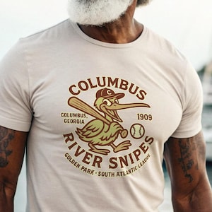 Puede incluir: Camiseta beige de cuello redondo con un gráfico de un pelícano jugando al béisbol y el texto "Columbus River Snipes" y "1909". El diseño es en tonos marrones y verdes. La camiseta la lleva una persona con pelo gris y barba.