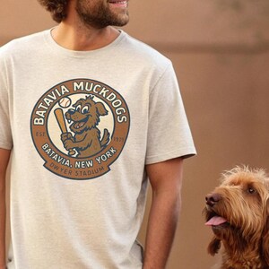 Puede incluir: Camiseta gris claro con un logotipo circular. El logotipo muestra el texto "Batavia Muckdogs" y un perro de dibujos animados con un bate de béisbol. La camiseta también incluye el texto "Batavia, New York" y "Dwyer Stadium".
