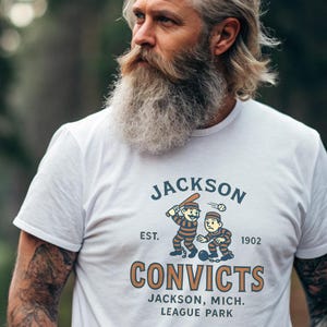 Op de afbeelding: Wit T-shirt met een afbeelding van twee cartoon honkbalspelers en de tekst "Jackson Convicts" in oranje en blauw. Het shirt bevat ook de tekst "Jackson, Mich. League Park."