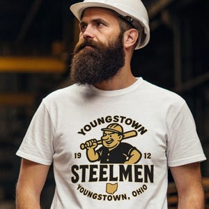 Puede incluir: Camiseta blanca con un jugador de béisbol de dibujos animados sosteniendo un bate, con el texto "YOUNGSTOWN STEELMEN" y "YOUNGSTOWN, OHIO". La camiseta también incluye los números "19" y "12". La camiseta la lleva una persona con un casco blanco.