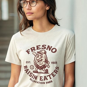 Puede incluir: Camiseta beige con un gráfico granate de un personaje de pasa jugando al béisbol. El texto en la camiseta dice "Fresno Raisin Eaters Recreation Park" y "Est. 1906". La camiseta tiene cuello redondo y mangas cortas.