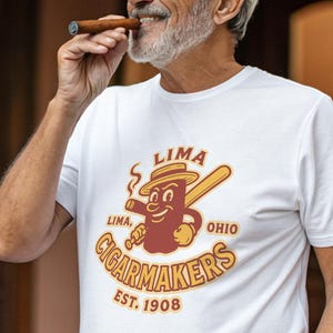 以下が含まれることがあります： 漫画の葉巻キャラクターと野球バットのデザインが特徴の白いTシャツで、「LIMA CIGARMAKERS OHIO EST. 1908」の文字が入っています。男性は葉巻を持っています。シャツは柔らかく快適な素材で作られています。