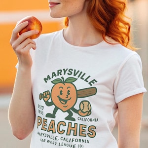 Puede incluir: Camiseta blanca con un diseño de melocotón de dibujos animados sosteniendo un bate de béisbol y el texto "Marysville Peaches, California". La camiseta tiene un diseño vintage, ideal para los fanáticos del béisbol.