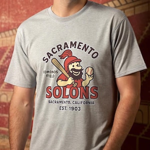 May include: A gray t-shirt featuring the Sacramento Solons baseball team logo. The logo includes a cartoon character with a baseball bat and ball, with text that reads "Sacramento Solons, Edmonds Field, Sacramento, California, Est. 1903."