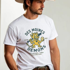 Puede incluir: Camiseta blanca con el logo del equipo de béisbol Des Moines Demons. El logo presenta un diablo amarillo con un bate y una pelota de béisbol. La camiseta también tiene el texto "Des Moines Demons" y "Holcomb Park".