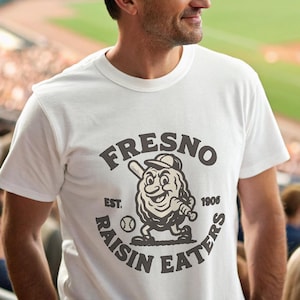Puede incluir: Camiseta blanca con el texto "Fresno Raisin Eaters" y un personaje de dibujos animados de una pasa con un bate de béisbol. La camiseta también tiene el texto "Est. 1906". Un hombre con una gorra de béisbol negra lleva la camiseta.