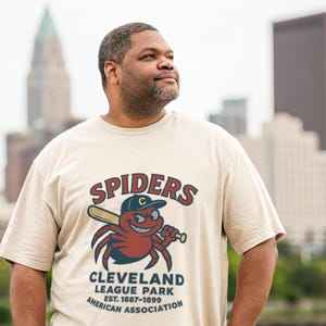 May include: Beige t-shirt with a cartoon spider holding a baseball bat. The text "SPIDERS" is above the spider, and "CLEVELAND LEAGUE PARK EST. 1887-1899 AMERICAN ASSOCIATION" is below. The spider is red with a blue baseball cap.