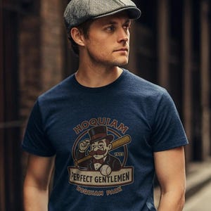 Può includere: T-shirt blu navy con grafica vintage. La grafica include il testo "HOQUIAM" sopra un'illustrazione di un gentiluomo con baffi, una mazza da baseball e le parole "PERFECT GENTLEMEN" e "HOQUIAM PARK".