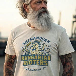 May include: A light beige t-shirt with a baseball-themed graphic. The design features a cartoon character with a baseball bat and the text "SHENANDOAH HUNGARIAN RIOTERS" in blue and yellow. The shirt also has the text "SHENANDOAH, PA".