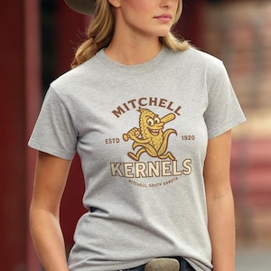 Könnte beinhalten: Graues T-Shirt mit einem Cartoon-Maiskolben, der einen Baseballschläger hält. Auf dem Shirt stehen die Worte "Mitchell Kernels" und "Mitchell, South Dakota". Gegründet 1920.