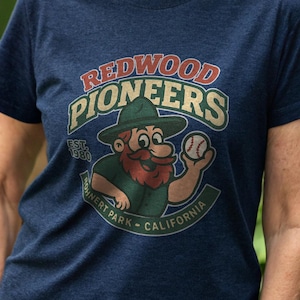 May include: Navy blue t-shirt with the text "Redwood Pioneers" in red and green. The graphic includes a cartoon character with a beard and baseball. The shirt is a crew neck and appears to be made of a soft material.