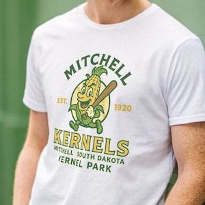 Könnte beinhalten: Weißes T-Shirt mit einem Cartoon-Maiskolben, der einen Baseballschläger hält. Das Shirt zeigt den Text "Mitchell Kernels" in Grün und Gelb, mit "Est. 1920". Die Person, die das Shirt trägt, hat rotes Haar.