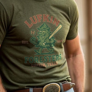 May include: Olive green t-shirt featuring the text "LUFKIN FORESTERS LUFKIN, TEXAS" and a cartoon tree with a baseball bat. The shirt is worn with a cowboy hat and a brown belt with a decorative buckle.