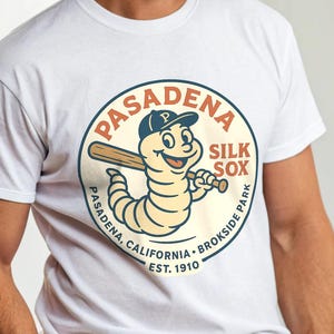 May include: White t-shirt with a circular graphic featuring a baseball-playing worm wearing a cap and holding a bat. The graphic reads "Pasadena Silk Sox" and "Pasadena, California, Brokside Park, Est. 1910". The person is wearing a beige cowboy hat.