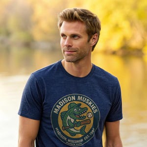 May include: A navy blue t-shirt featuring the "Madison Muskies" logo. The logo depicts a cartoon fish wearing a baseball cap and holding a bat. The text "Madison, Wisconsin, Warner Park, Est. 1982" is also included.