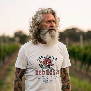 May include: White t-shirt with the text "LANCASTER RED ROSES" and a baseball-themed graphic. The graphic includes a smiling red rose, baseball bat, and baseball. Additional text includes "LANCASTER, PA TRI-STATE LEAGUE STUMPF FIELD EST. 1906".