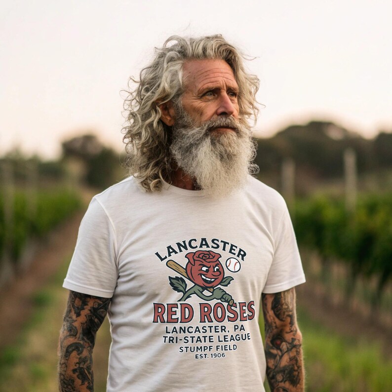 May include: White t-shirt with the text "LANCASTER RED ROSES" and a baseball-themed graphic. The graphic includes a smiling red rose, baseball bat, and baseball. Additional text includes "LANCASTER, PA TRI-STATE LEAGUE STUMPF FIELD EST. 1906".