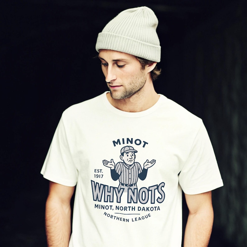 Op de afbeelding: Cr&egrave;mekleurig T-shirt met een honkbalthema-afbeelding met de tekst "MINOT WHY NOTS" in marineblauw. Het shirt heeft ook de tekst "MINOT, NORTH DAKOTA NORTHERN LEAGUE" en "EST. 1917". Een lichtgrijze beanie is ook zichtbaar.