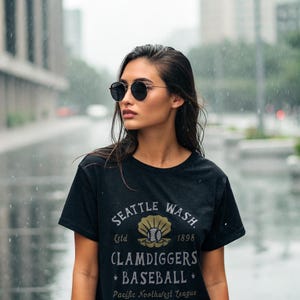 May include: Black t-shirt with the text "SEATTLE WASH CLAMDIGGERS BASEBALL" in a vintage design. The shirt features a baseball graphic and the text "Pacific Northwest League". The person is wearing round black sunglasses.