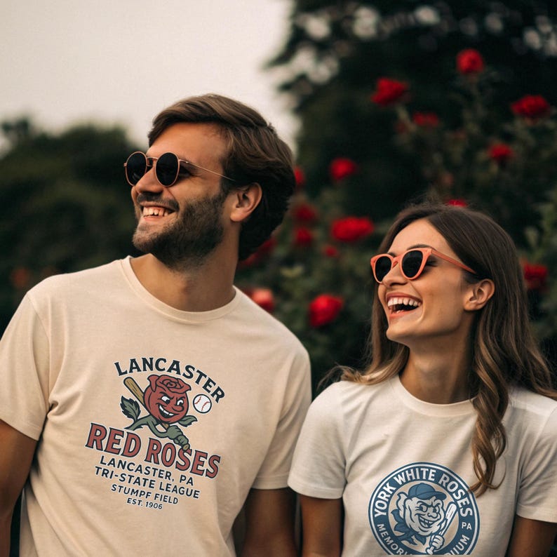 May include: Two people wearing sunglasses and smiling, each wearing a t-shirt. One shirt says "Lancaster Red Roses" with a baseball and rose graphic. The other shirt says "York White Roses" with a baseball graphic.
