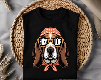 May include: Black t-shirt featuring a cartoon basset hound wearing sunglasses with the text "Rocky Top". The dog has a checkered bandana and head covering. The shirt is displayed on a woven basket.
