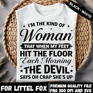 Puede incluir: Camiseta blanca con una cita humorística en texto negro: "I'M THE KIND OF Woman THAT WHEN MY FEET HIT THE FLOOR Each Morning THE DEVIL SAYS OH CRAP SHE'S UP." La camiseta se muestra en una cesta tejida.