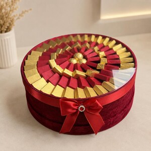 May include: A round, red velvet gift box with a clear lid, filled with rectangular chocolates wrapped in gold and red foil. The box is decorated with a red satin ribbon bow featuring a central rhinestone accent, arranged in a circular pattern.