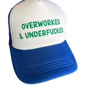 May include: A blue and white trucker hat with the text "OVERWORKED & UNDERFUCKED" in green. The hat has a blue brim and mesh back, with a white front panel. The hat features a curved brim and a button on top.