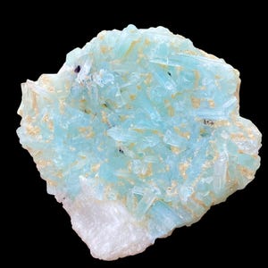Puede incluir: Primer plano de un espécimen mineral con estructuras cristalinas de color azul claro. Los cristales se mezclan con áreas de color beige y descansan sobre una base blanca, sobre un fondo negro. La forma es irregular y texturizada. Es una formación natural.