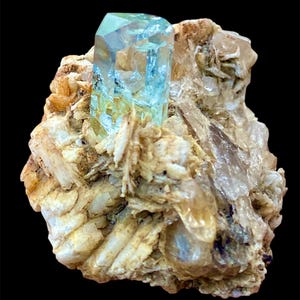 May include: A striking aquamarine crystal, with a clear, light blue hue, is the focal point of this image. It is set within a matrix of beige and tan minerals, creating a natural, earthy contrast. The crystal's facets catch the light, showcasing its crystalline structure.