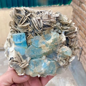 May include: A natural aquamarine crystal cluster with a striking blue hue. The crystal is surrounded by a matrix of beige, layered mineral formations. The rough, uncut gemstone is a beautiful example of natural mineral formations.