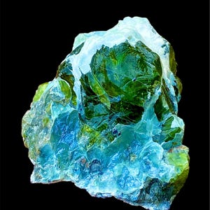 Puede incluir: Primer plano de un espécimen mineral sobre un fondo oscuro. El mineral presenta una estructura cristalina con tonos verdes, azules y blancos. Los colores se mezclan, creando una apariencia translúcida. La superficie del mineral es rugosa y desigual.