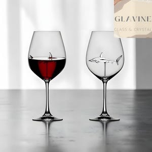 May include: Two clear glass wine glasses with long stems. One glass is filled with red wine and contains a small shark figure with only its dorsal fin visible. The other empty glass features a full silver-toned shark figure inside the bowl.