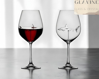 May include: Two clear glass wine glasses with long stems. One glass is filled with red wine and contains a small shark figure with only its dorsal fin visible. The other empty glass features a full silver-toned shark figure inside the bowl.