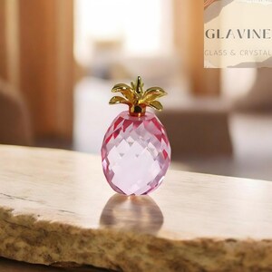 Peut inclure: Une petite figurine d'ananas en cristal rose à facettes avec un sommet en métal doré poli. Le corps de l'ananas présente un motif géométrique en diamant qui reflète la lumière, tandis que les feuilles métalliques à la couronne sont lisses.