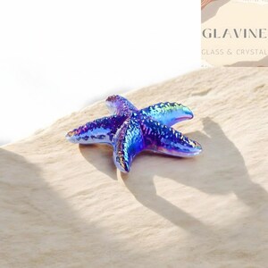 May include: A five-pointed starfish figurine featuring a vibrant, iridescent finish that shifts between deep blue, purple, and teal tones. The surface has a textured, bumpy appearance with a glossy, reflective sheen throughout the entire object.