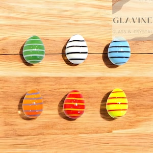 May include: Six colorful glass Easter egg ornaments with horizontal stripes in green, black and white, blue, orange and purple, red and yellow, and yellow and red. The eggs are on a wooden surface. The text "GLAVINE GLASS & CRYSTAL" is in the upper right corner.