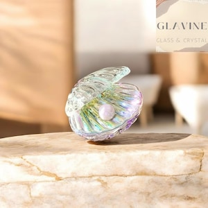 May include: A decorative glass seashell ornament featuring an iridescent finish with a soft purple, green, and blue sheen. The shell is partially open, revealing a single, smooth, round, light-coloured pearl nestled inside the centre of the glass structure.
