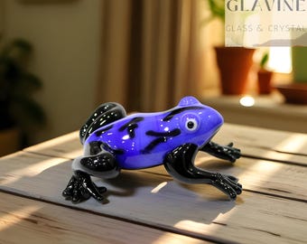Könnte beinhalten: Eine Glasfroschfigur mit einem leuchtend blauen Körper und schwarzen Akzenten. Die Augen des Frosches sind weiß, die Beine schwarz. Die Figur steht auf einer Holzoberfläche. Der Text "GLAVINE GLASS & CRYSTAL" ist ebenfalls zu sehen.