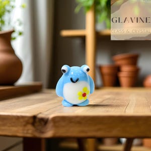 以下が含まれることがあります： 白腹と黄色の花の模様が付いた、水色のカエルの置物。カエルは大きな黒い目と笑顔の口をしています。置物はガラスまたはクリスタル製で、木製の表面に置かれています。