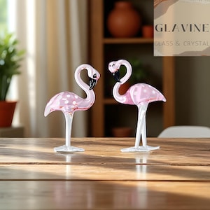 May include: Two glass flamingo ornaments. The flamingos are pink with white polka dots and black beaks. They stand on clear glass legs and bases. The ornaments are displayed on a wooden table.