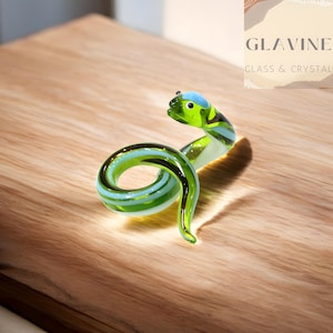 May include: A glass snake ornament in shades of green and blue, coiled on a wooden surface. The snake has a small head and is made of glass. The brand name "GLAVINE GLASS & CRYSTAL" is visible in the background. A decorative item.