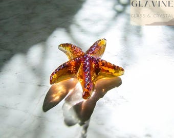 May include: A translucent amber and gold glass starfish figurine with a textured, bumpy surface. The object features a five-pointed star shape with a glossy finish that catches the light, creating a warm, reflective glow across its surface.