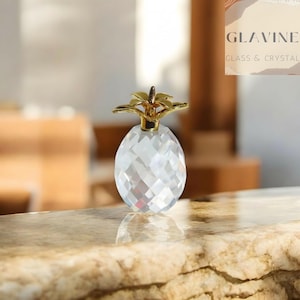 May include: A clear, faceted crystal glass figurine shaped like a pineapple with a polished gold-tone metal top. The object features a geometric, diamond-cut pattern across its rounded body and a detailed, multi-pointed leaf design at the crown.