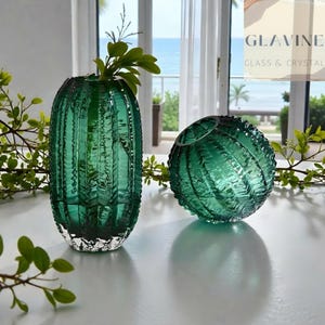 Peut inclure: Deux vases en verre vert émeraude avec une surface texturée et nervurée rappelant un cactus. Un vase est grand et cylindrique, tandis que l'autre est rond et sphérique. Le texte GLAVINE GLASS & CRYSTAL est visible dans le coin supérieur droit.