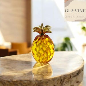 Może przedstawiać: Fasetowana figurka ananasa ze szkła w kolorze bursztynowym z polerowaną, złotą metalową górą. Ananas posiada geometryczny wzór w romby na całym korpusie oraz odblaskową powierzchnię, z widoczną nazwą marki GLAVINE GLASS & CRYSTAL.