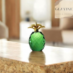 May include: A decorative pineapple figurine crafted from faceted green glass with a polished gold-tone metal top. The object features a geometric diamond-cut pattern across the body and a stylised leaf crown, reflecting light through its translucent surface.