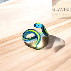 May include: A glass snake ornament in shades of blue, yellow, and green. The coiled snake has a detailed head and is set on a light wood surface. The background features the text "GLAVINE GLASS & CRYSTAL".