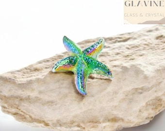 May include: A small, iridescent glass starfish figurine featuring a vibrant spectrum of green, blue, and purple hues with a textured surface. The object is positioned on a rough, light-colored stone. The text GLAVINE GLASS & CRYSTAL is visible.